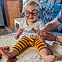 baby, child, glasses, striped_pants, broomstick, blanket, smile, indoor, person, hand, floor, wooden_furniture, couch, decor, plaid_blanket, foot, toddler, cute, costume, happy