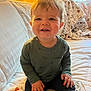 toddler, child, smiling, indoor, couch, blanket, long_sleeve_shirt, pants, hands, face, hair, lamp, wood_paneling, window, curtains, fabric, cozy, home, seated, portrait