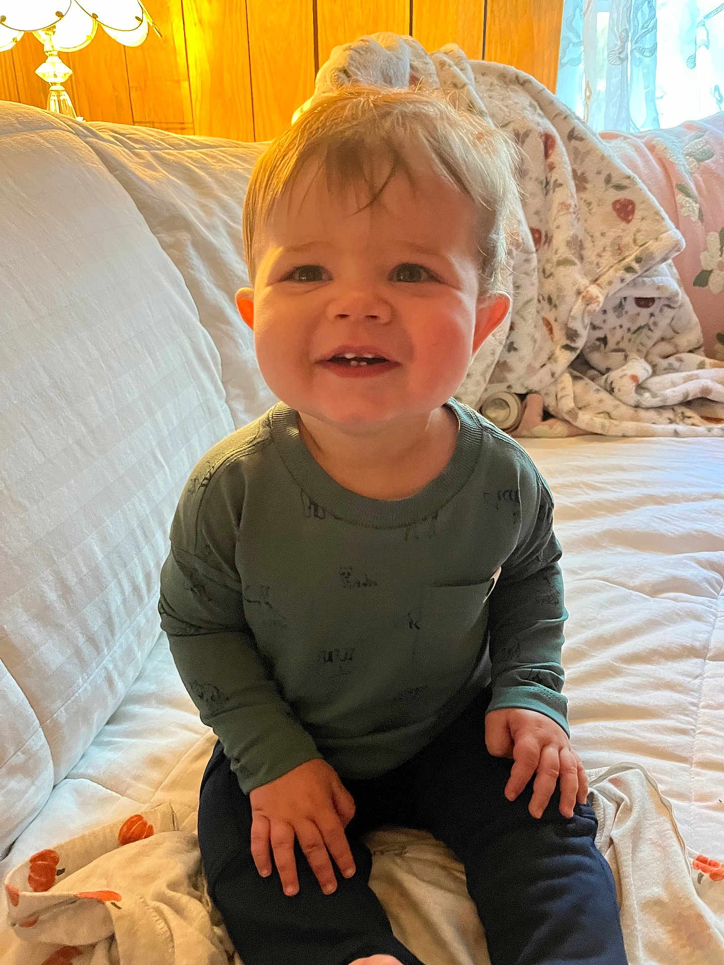 George is registered to the contest to win money with this photo: toddler, child, smiling, indoor, couch, blanket, long_sleeve_shirt, pants, hands, face, hair, lamp, wood_paneling, window, curtains, fabric, cozy, home, seated, portrait