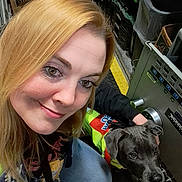 Courage is registered to the contest to win money with this photo: woman, puppy, dog, selfie, smiling, portrait, face, blonde_hair, blue_eyes, gray_coat, safety_vest, workshop, machinery, industrial, equipment, storage_bins, indoor, close_up, sitting, photography