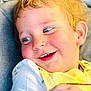 baby, blonde_hair, blue_eyes, casual_clothing, child, closeup, cute, expression, face, happy, indoors, joyful, person, portrait, seated, smiling, soft_lighting, toddler, white_shirt, yellow_bib