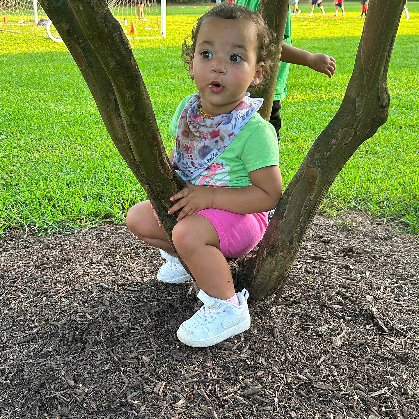 Cassia is registered to the contest to win money with this photo: background, bandana, child, curious, daytime, expression, field, grass, greenery, nature, outdoor, people, pink_shorts, playing, sneakers, soccer, sports, sunlight, toddler, tree
