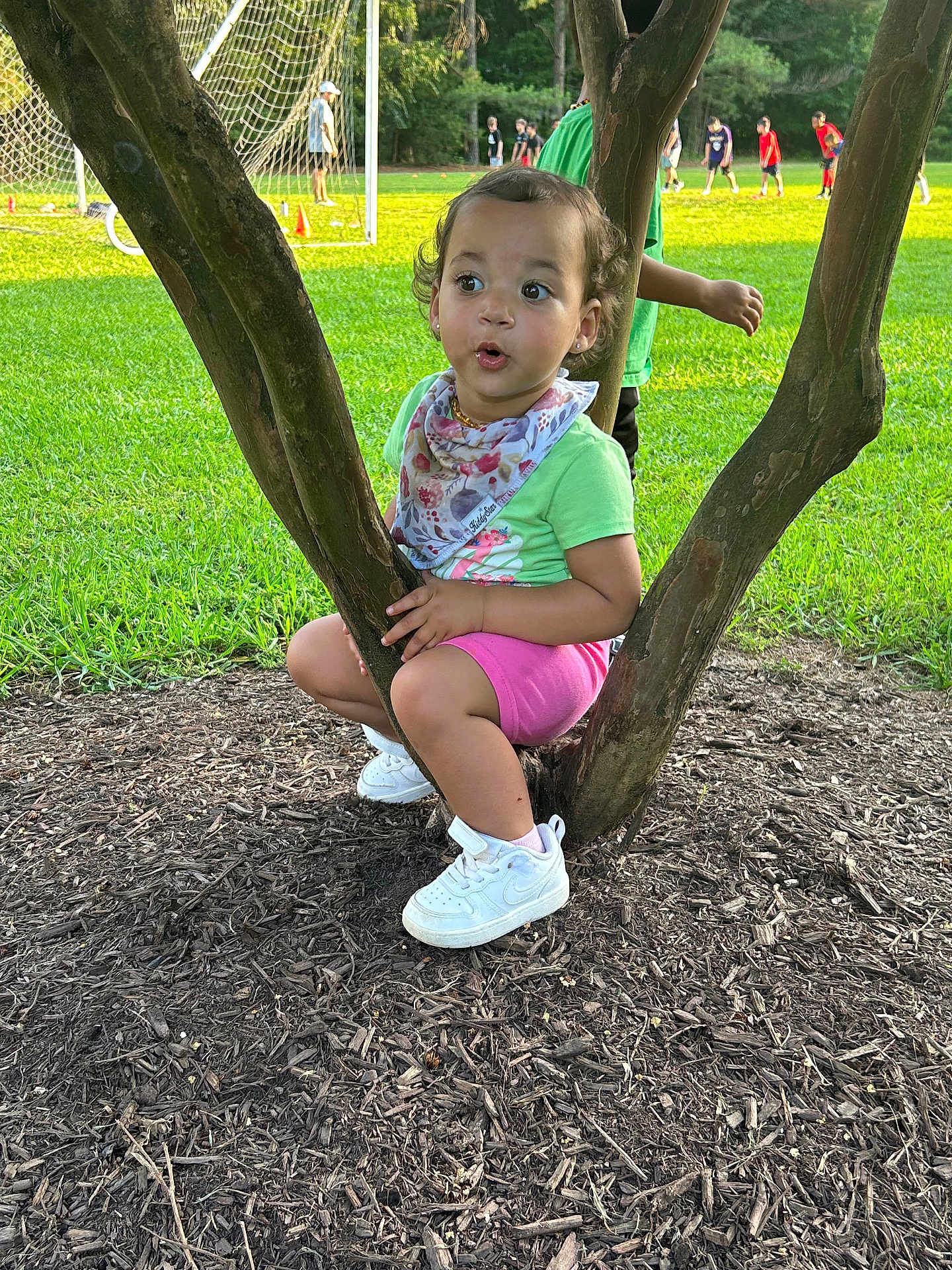 Cassia is registered to the contest to win money with this photo: background, bandana, child, curious, daytime, expression, field, grass, greenery, nature, outdoor, people, pink_shorts, playing, sneakers, soccer, sports, sunlight, toddler, tree