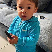 Zackaria a rejoint le concours — aidez-le/la à gagner de superbes lots ! black_shoes, blue_tracksuit, casual_clothing, child, couch, curious, cushions, expression, face, floor_tiles, home, indoor, living_room, nike_logo, person, portrait, remote_control, standing, toddler, young_child