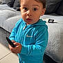 black_shoes, blue_tracksuit, casual_clothing, child, couch, curious, cushions, expression, face, floor_tiles, home, indoor, living_room, nike_logo, person, portrait, remote_control, standing, toddler, young_child