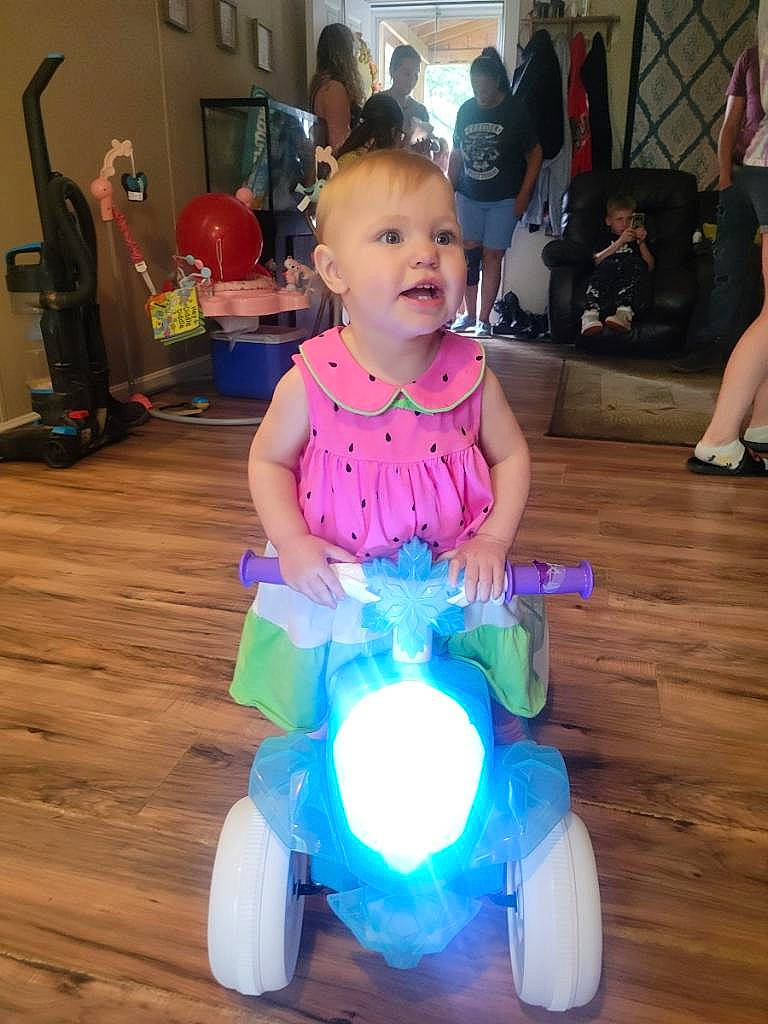 Scarlette is registered to the contest to win money with this photo: baby, baby_toddler_clothing, child, electric_blue, event, flooring, fun, happy, hardwood, leisure, magenta, person, purple, riding_toy, shoe, sitting, smile, television, toddler, toy