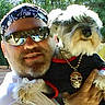man, dog, outdoor, sunglasses, bandana, pet, holding, black_clothing, necklace, skull, fence, greenery, sunlight, portrait, beard, casual, happy, small_dog, yard, relaxed