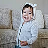 toddler, child, smile, hoodie, white_clothing, couch, beige, snack, happy, indoor, cozy, plush_pillow, knitwear, cute, person, young_child, casual_wear, home, portrait, standing