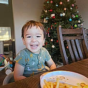 Theodore is registered to the contest to win money with this photo: child, toddler, smile, happy, indoor, christmas_tree, ornaments, holiday, table, plate, french_fries, chair, wooden_furniture, clothing, short_sleeve, face, person, home, dining, toy