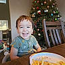 child, toddler, smile, happy, indoor, christmas_tree, ornaments, holiday, table, plate, french_fries, chair, wooden_furniture, clothing, short_sleeve, face, person, home, dining, toy