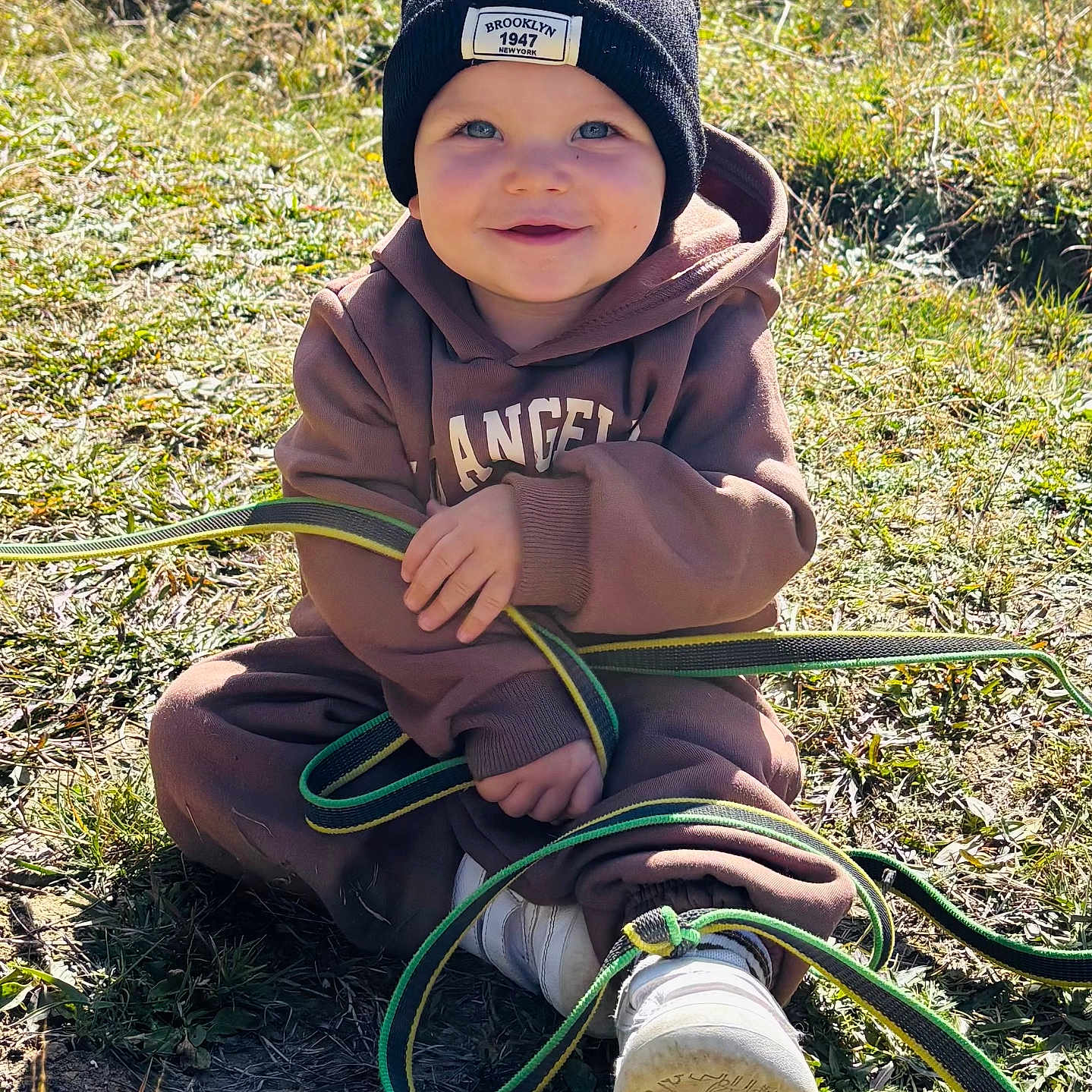 Maélïo a rejoint le concours — aidez-le/la à gagner de superbes lots ! beanie, black_hat, brown_clothing, casual, child, cute, daylight, grass, happy, hoodie, nature, outdoor, playful, shoes, sitting, smiling, strap, sunlight, toddler, young_child