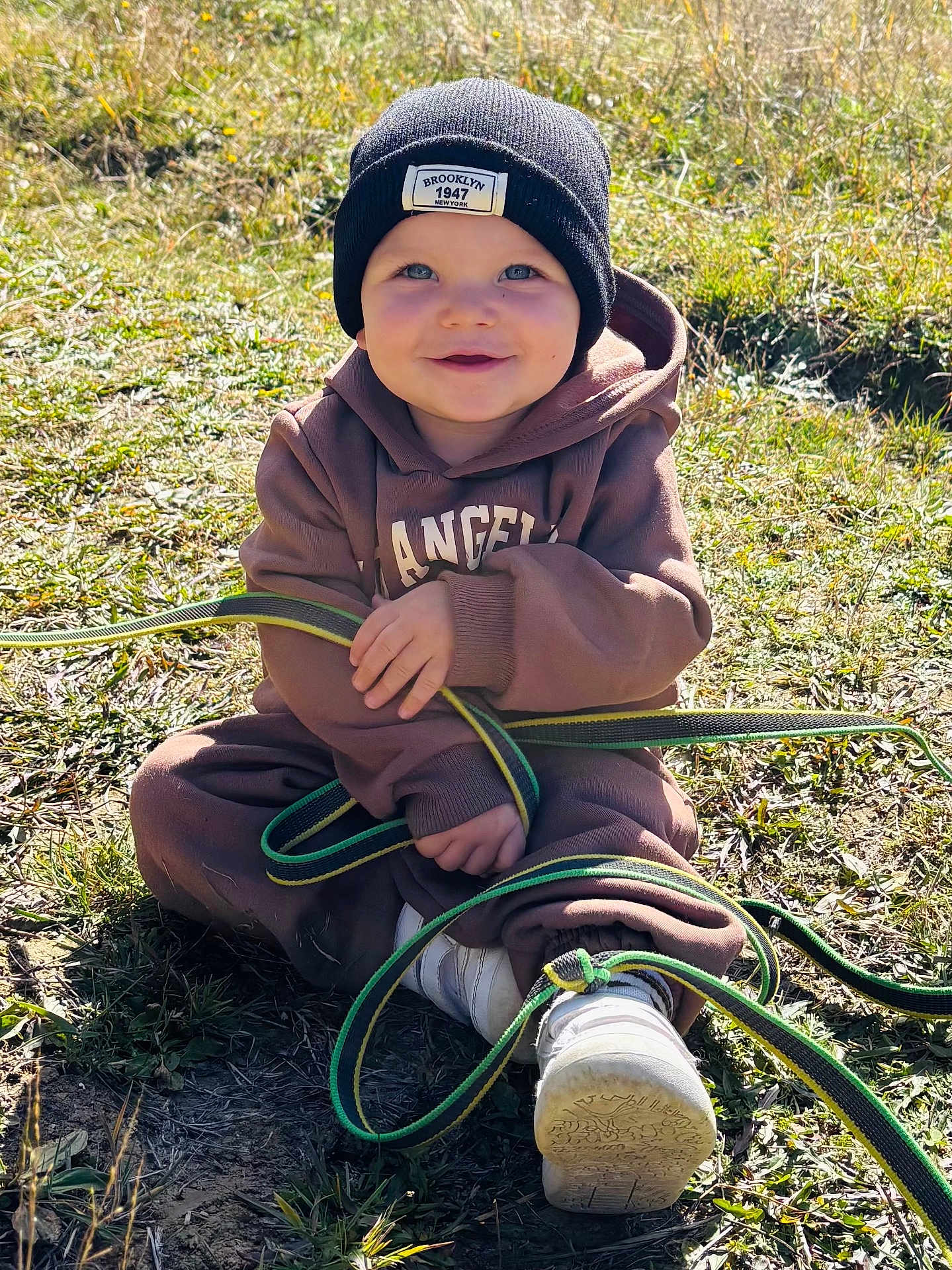 Maélïo a rejoint le concours — aidez-le/la à gagner de superbes lots ! toddler, child, grass, outdoor, sunlight, beanie, black_hat, hoodie, brown_clothing, smiling, happy, playful, strap, shoes, casual, nature, daylight, sitting, cute, young_child