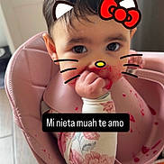 Penelope joined the competition — help win amazing prizes! baby, infant, child, high_chair, sitting, portrait, face, eyes, dark_hair, smiling, hand, food, food_splatter, pink, bib, floral_clothing, kitchen, indoors, hello_kitty_filter, text_overlay