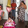 child, toddler, birthday, party, toy_car, pink, balloon, number_one, family, adults, indoor, celebration, streamers, dress, sitting, smiling, home, wood_floor, decorations, portrait