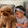 baby, child, dog, poodle, curly_hair, bed, bedroom, headboard, pajamas, dress, headband, pink_socks, sitting, pet, companion, indoors, portrait, fluffy, smiling, family