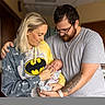 baby, newborn, adult, woman, man, batman_hoodie, tattoo, glasses, indoor, family, love, parent, child, holding, smiling, casual_clothing, portrait, tenderness, affection, people
