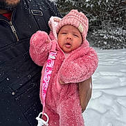 Billie is registered to the contest to win money with this photo: baby, infant, child, winter_clothing, pink_suit, knit_hat, pacifier_clip, snow, snowfall, adult_hand, outdoor, cold_weather, warm_clothing, face, person, coat, jacket, fuzzy_texture, nature, tree