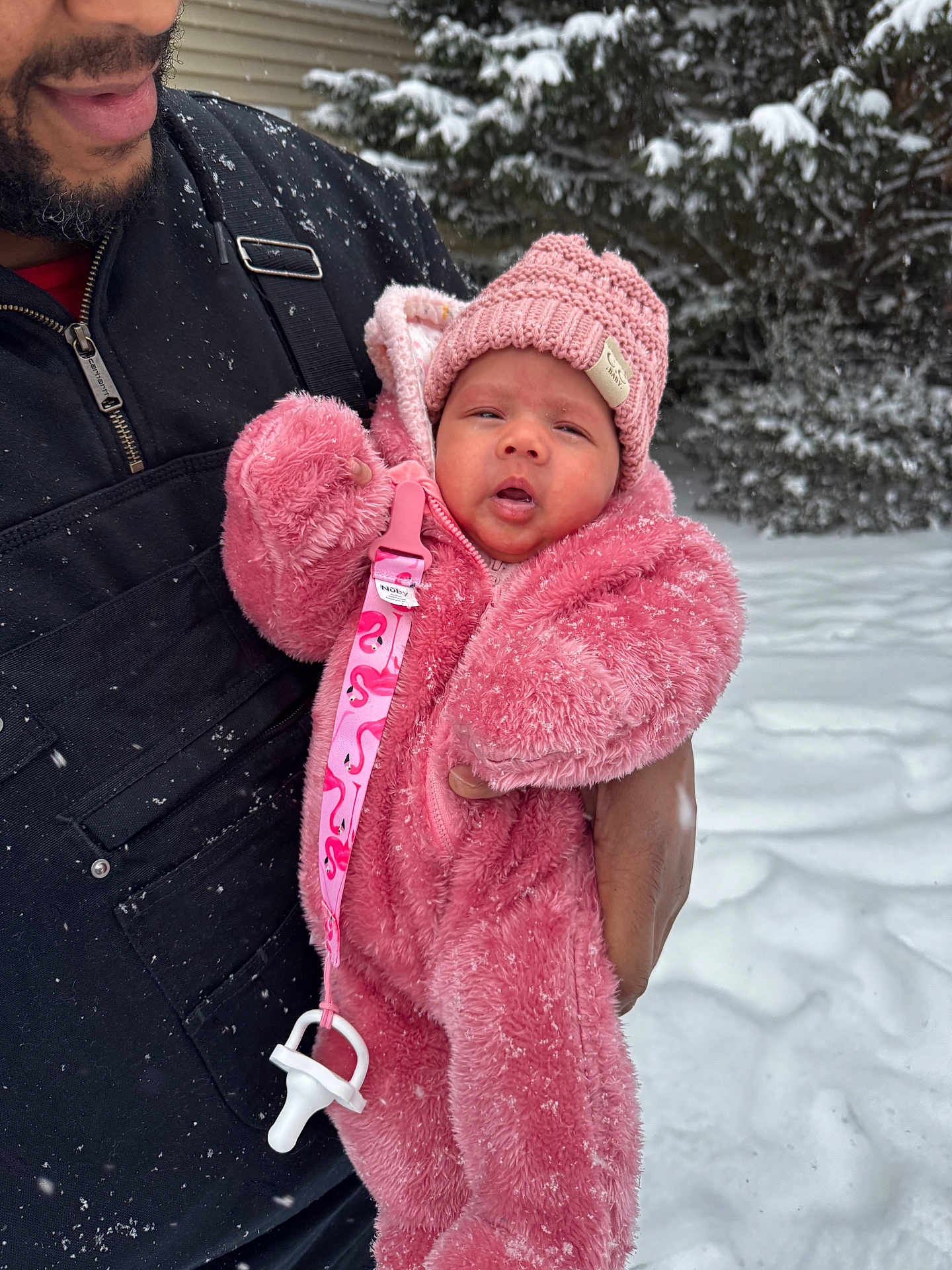 Billie is registered to the contest to win money with this photo: baby, infant, child, winter_clothing, pink_suit, knit_hat, pacifier_clip, snow, snowfall, adult_hand, outdoor, cold_weather, warm_clothing, face, person, coat, jacket, fuzzy_texture, nature, tree