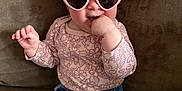 Coralie is registered to the contest to win money with this photo: baby, child, pink_headband, heart_sunglasses, couch, hand, blue_jeans, patterned_shirt, smiling, indoors, cute, playful, sitting, finger_in_mouth, headwear, fashion, person, comfortable, cozy, tickling