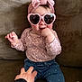 baby, child, pink_headband, heart_sunglasses, couch, hand, blue_jeans, patterned_shirt, smiling, indoors, cute, playful, sitting, finger_in_mouth, headwear, fashion, person, comfortable, cozy, tickling