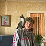 animal, casual_clothing, cat, curtain, decor, facial_hair, framed_picture, furniture, hair, hoodie, indoor, man, pet, plant, portrait, potted_plant, television, wall, window, young_adult