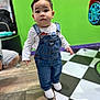 toddler, child, denim_overalls, white_shirt, white_shoes, indoors, checkered_floor, green_wall, curious, standing, person, short_hair, flooring, clothing, casual, portrait, young_child, cute, looking_at_camera, play_area