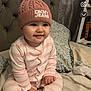 baby, child, infant, hat, knit_hat, pom_pom, pink, striped_clothing, onesie, bed, pillow, headboard, smiling, indoors, cozy, cute, person, face, sitting, sleepwear