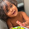 child, toddler, smiling, portrait, curly_hair, brown_hair, face, teeth, eyes, indoor, close_up, sitting, couch, bare_shoulders, toy, ball, colorful, happy, candid, photography