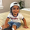 child, toddler, portrait, smiling, sitting, bedroom, bed, baseball_cap, backward_cap, wet_hair, t_shirt, jeans, shorts, sneakers, socks, indoor, home, bedding, plant, fashion