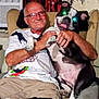 Booboo is registered to the contest to win money with this photo: adult, air_conditioner, black_and_white_dog, cartoon, casual_clothing, chair, dog, furniture, glasses, hand, happy, home, indoor, man, pet, seated, shirt, smile, tattoo, window