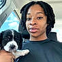 person, puppy, dog, car, selfie, twist_hairstyle, black_hair, white_puppy_paw, window, sky, clouds, seat, casual_clothing, hand, face, portrait, indoor, daylight, calm, cute