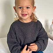 Naël a rejoint le concours — aidez-le/la à gagner de superbes lots ! toddler, child, smiling, curly_hair, blonde, dark_sweater, blue_jeans, sitting, indoor, white_background, small_object, happy, cute, portrait, casual_clothing, hands, young_child, person, light_skin, minimalist