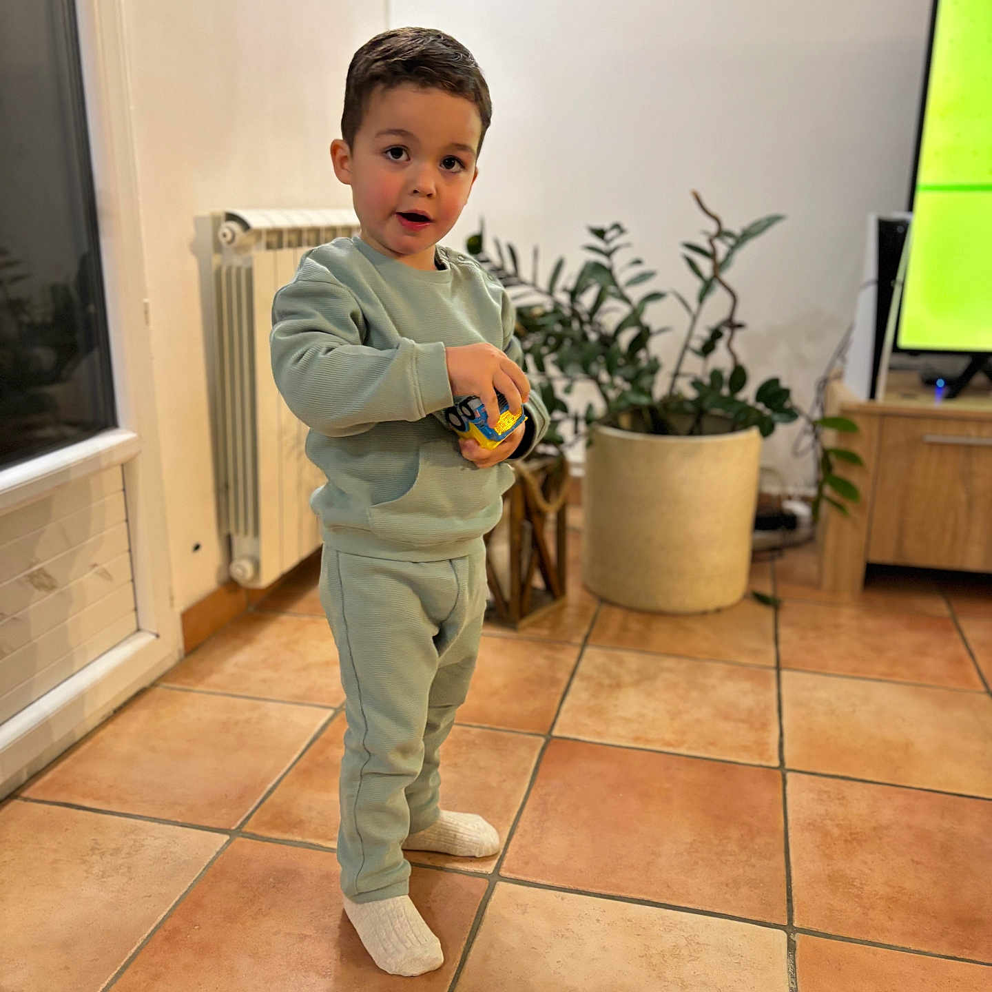 Noah a rejoint le concours — aidez-le/la à gagner de superbes lots ! casual, child, clothing, curious, expression, floor, furniture, home, indoor, living_room, person, plant, potted_plant, radiator, socks, standing, television, tile_floor, toddler, toy
