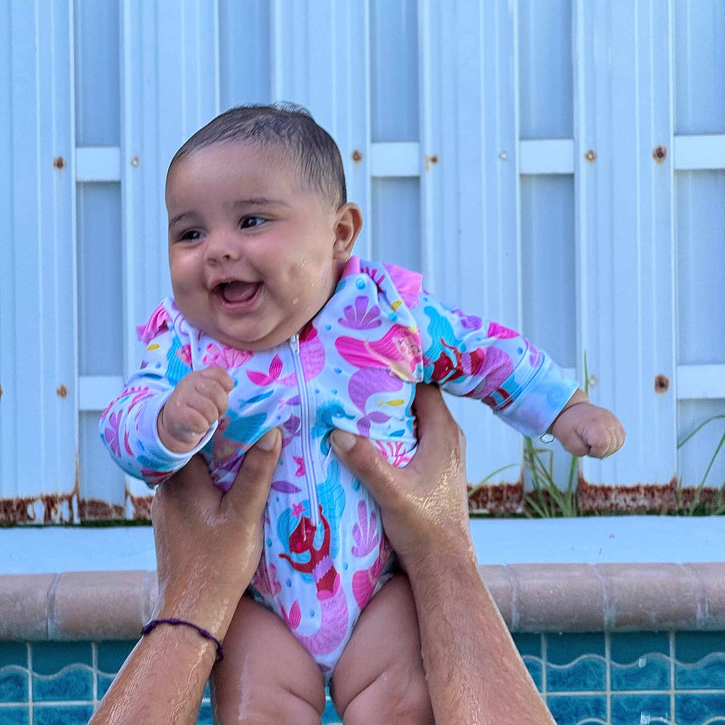 Amelia joined the competition — help win amazing prizes! baby, child, smiling, happy, portrait, outdoor, swimming_pool, water, held, infant, colorful_clothing, floral_pattern, onesie, hands, adult_hands, fence, backyard, summer, poolside, close_up