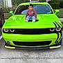 Kiannsha is registered to the contest to win money with this photo: toddler, child, car, green_car, hood, sitting, outdoor, suburban, parking_lot, clothing, sweater, jeans, shoes, hair_bow, face, vehicle, headlight, concrete, sidewalk, nature