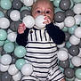 baby, child, ball_pit, plastic_balls, striped_clothing, romper, indoor, play, cute, infant, hand, face, eyes, legs, toy, colorful, soft_lighting, casual, smiling, fun