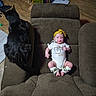 baby, black_cat, bow, cat, chair, child, furniture, headband, home, indoors, infant, living_room, lying_down, onesie, pacifier, pet, recliner, sitting, socks, toy