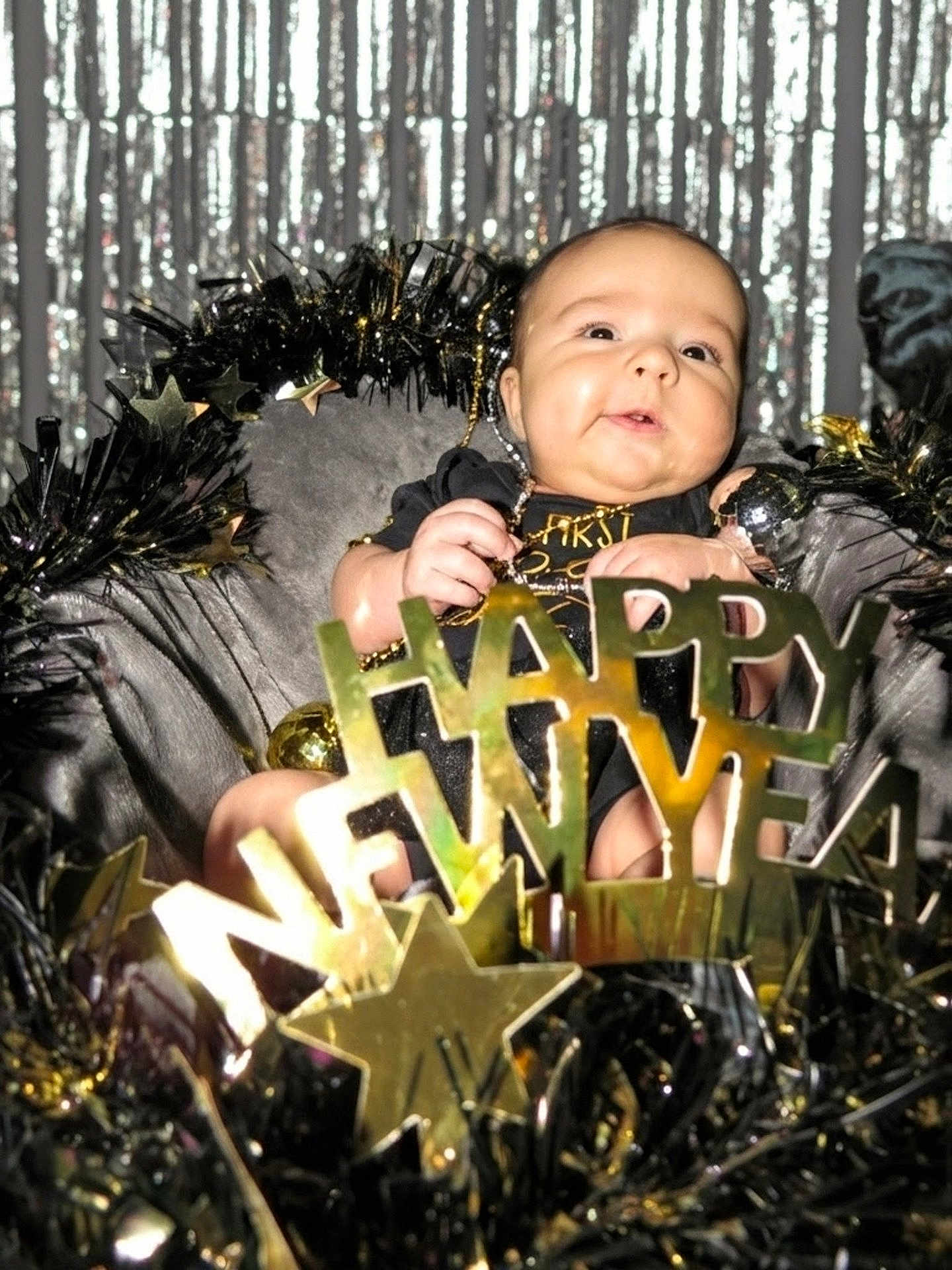 Hollie is registered to the contest to win money with this photo: baby, infant, new_year, celebration, gold, stars, tinsel, black_clothing, decorations, smiling, face, holiday, festive, sparkle, cute, portrait, background, party, child, holiday_decor