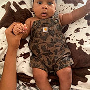 Blake is registered to the contest to win money with this photo: baby, child, infant, camo_overalls, blanket, cow_print, hand, holding, wide_eyes, expression, cute, lying_down, person, skin, face, fingers, legs, bed, comfort, soft