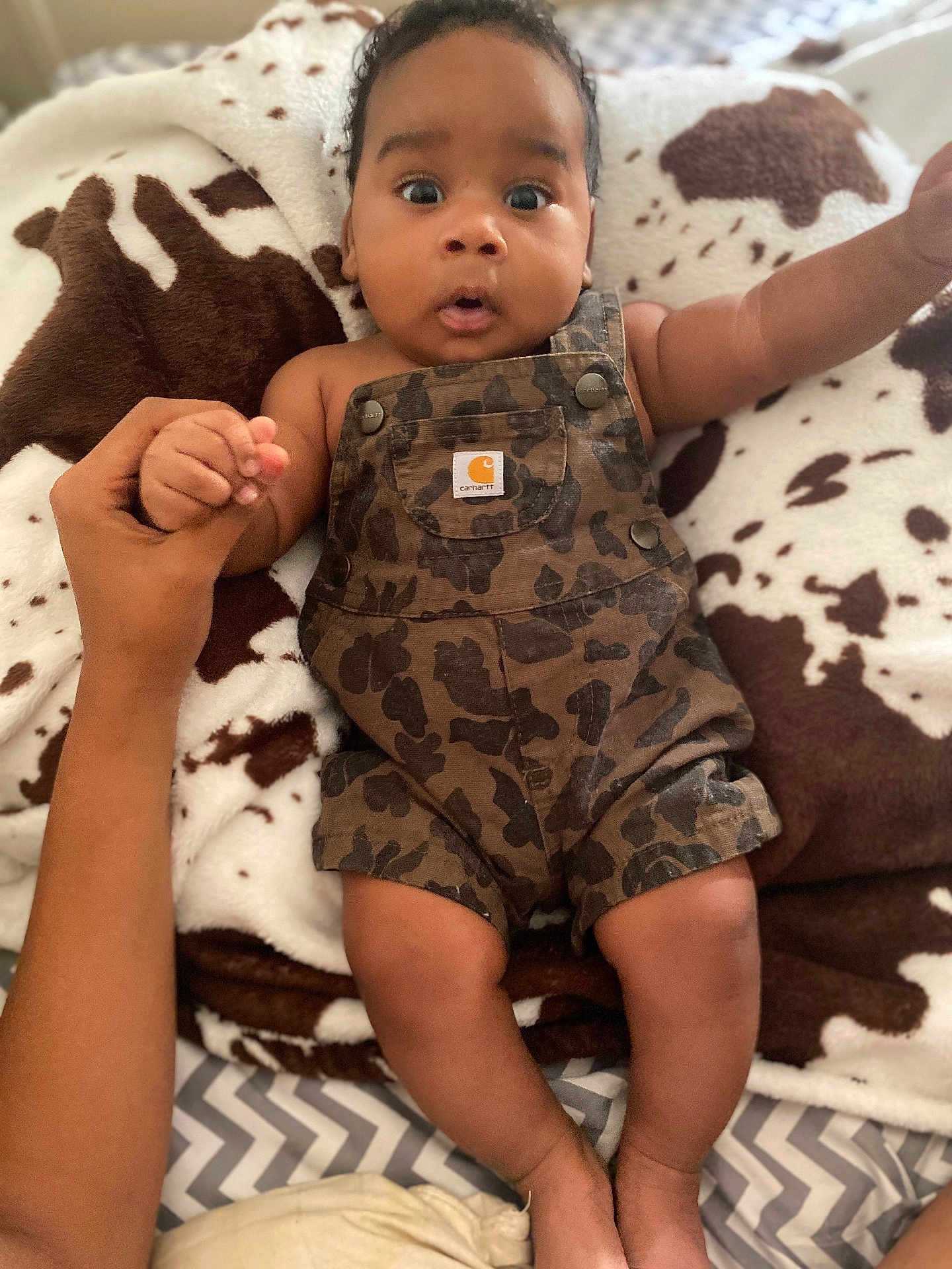 Blake is registered to the contest to win money with this photo: baby, child, infant, camo_overalls, blanket, cow_print, hand, holding, wide_eyes, expression, cute, lying_down, person, skin, face, fingers, legs, bed, comfort, soft