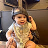 adult_hand, baby, backwards_cap, barefoot, booth, child, clothing, framed_picture, gray_onesie, happy, indoor, infant, portrait, restaurant, restaurant_interior, sitting, smiling, table, television, wood