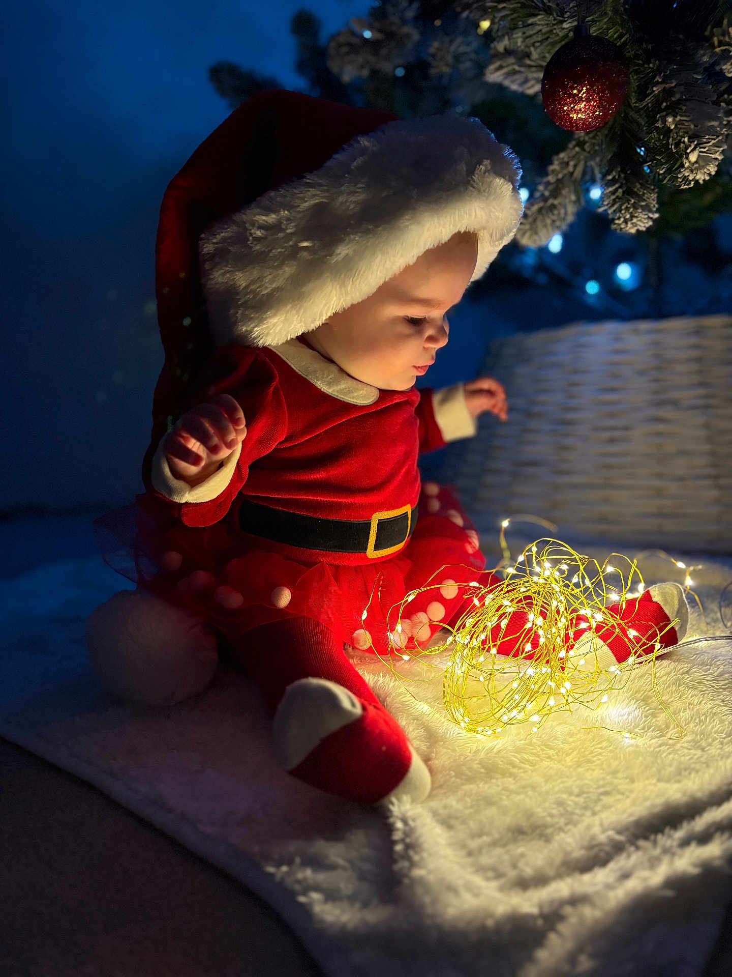 Harley Rose joined the competition — help win amazing prizes! baby, christmas, santa_hat, red_clothing, fairy_lights, blanket, holiday, decorations, tree, indoors, child, festive, cute, toddler, warm_lighting, cozy, celebration, smiling, face, sitting