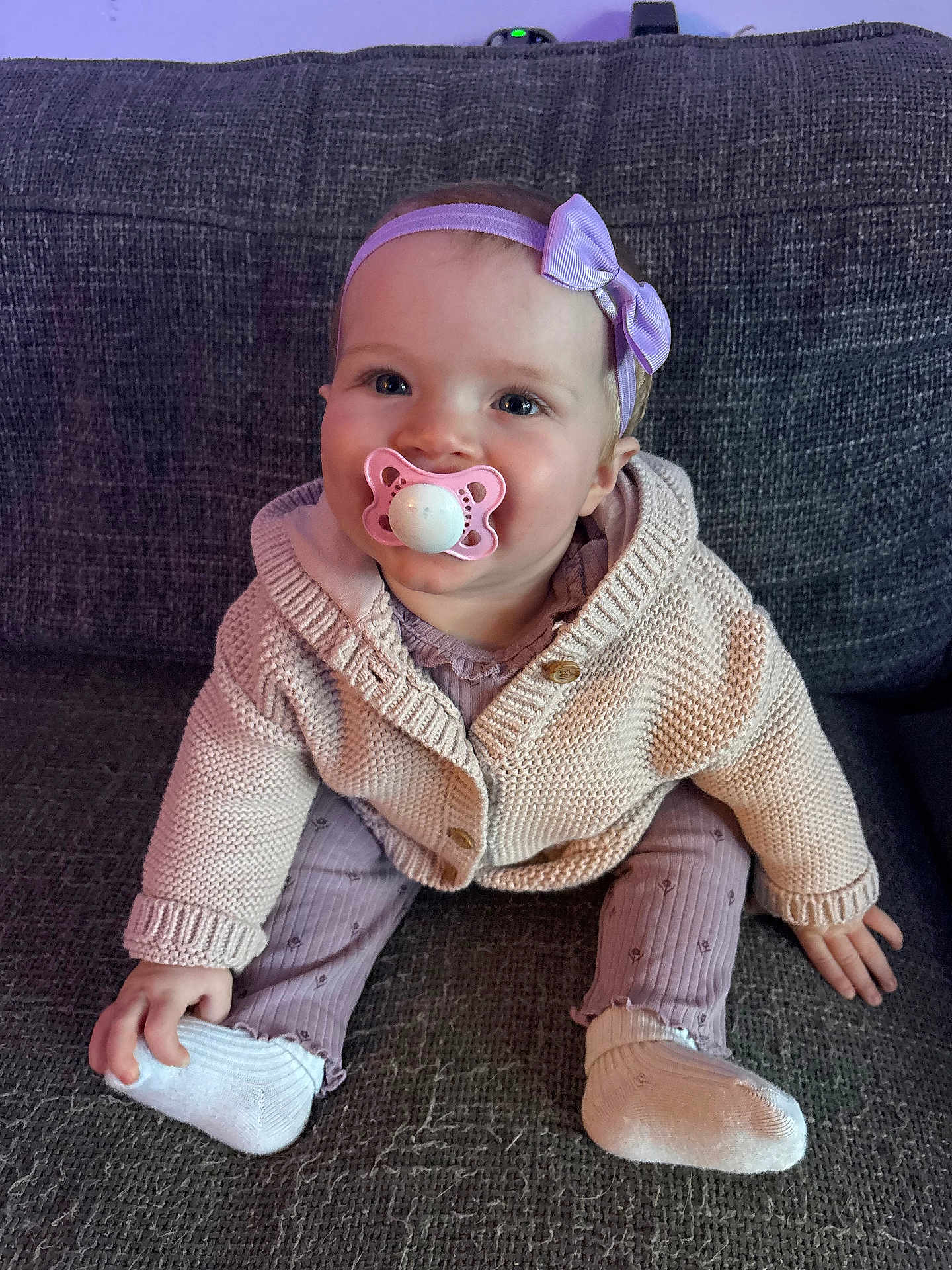 Harley Rose is registered to the contest to win money with this photo: baby, cardigan, child, clothing, couch, cute, footwear, happy, headband, indoor, infant, knitwear, pacifier, person, portrait, purple, sitting, smiling, sock, young_child