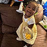 Daionna Rampey joined the competition — help win amazing prizes! toddler, child, smiling, yellow_dress, white_shirt, couch, snack_container, hair_beads, happy, indoor, brown_couch, clothing, footwear, crocs, toy, blanket, bag, person, portrait, home