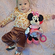 Elora is registered to the contest to win money with this photo: baby, child, blanket, plush_toy, minnie_mouse, headband, bow, floral_clothing, velvet_pants, socks, toy, floor, wooden_floor, smile, cute, lying_down, indoor, playful, soft_texture, portrait