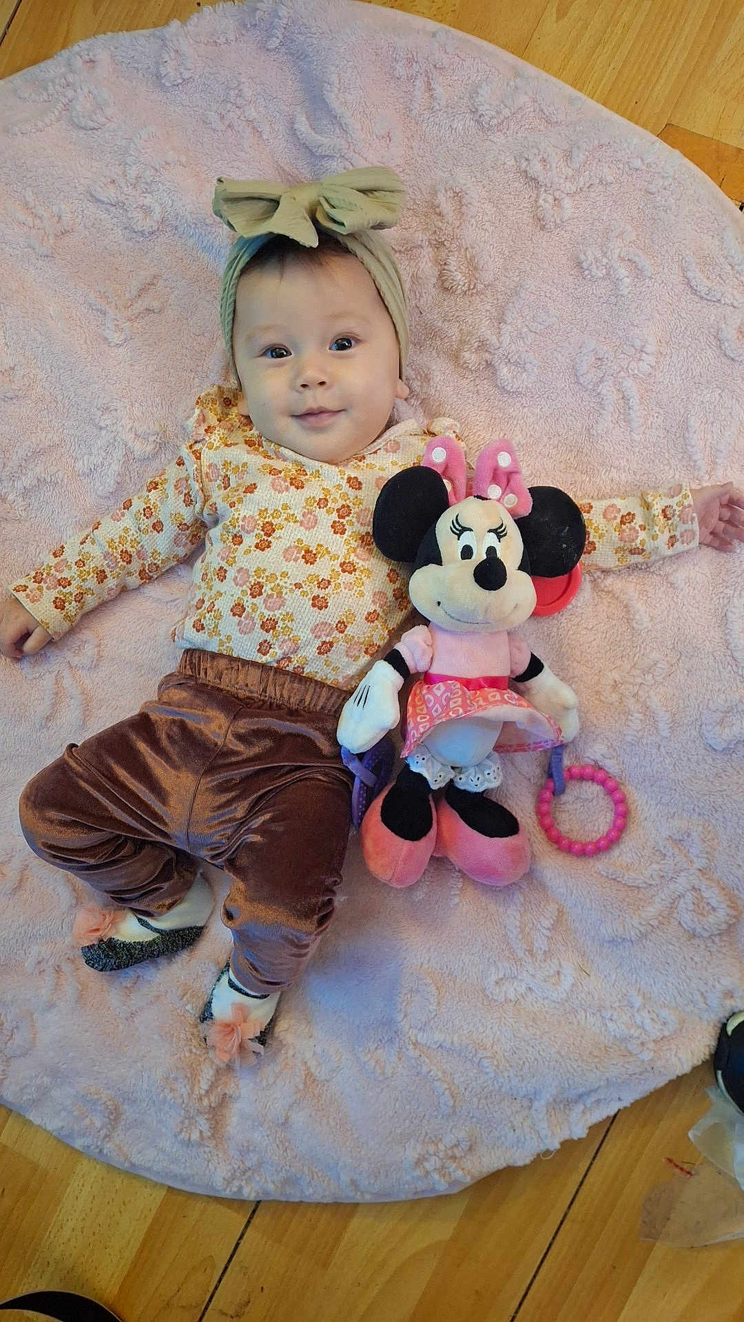 Elora is registered to the contest to win money with this photo: baby, child, blanket, plush_toy, minnie_mouse, headband, bow, floral_clothing, velvet_pants, socks, toy, floor, wooden_floor, smile, cute, lying_down, indoor, playful, soft_texture, portrait