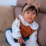 Laïa a rejoint le concours — aidez-le/la à gagner de superbes lots ! baby, child, headband, bow, couch, tights, white, brown, curly_hair, cute, smiling, portrait, indoor, cozy, clothing, sitting, curious, soft_lighting, floral, cushion