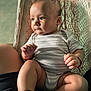 baby, infant, child, striped_onesie, hammock, sitting, legs, hands, skin, face, expression, natural_light, comfort, home, person, footwear, black_socks, red_bracelet, closeup, portrait