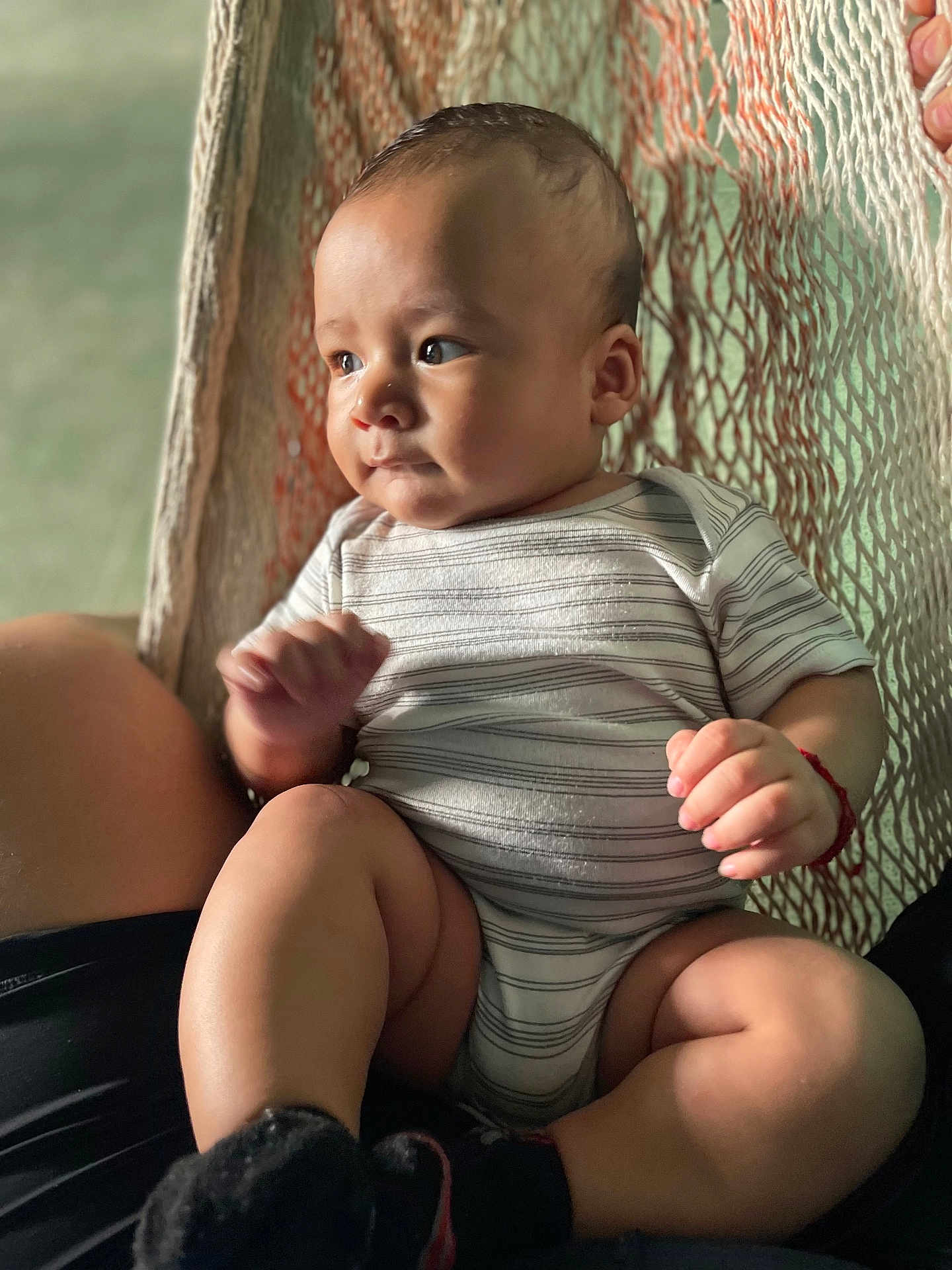 Beckham is registered to the contest to win money with this photo: baby, infant, child, striped_onesie, hammock, sitting, legs, hands, skin, face, expression, natural_light, comfort, home, person, footwear, black_socks, red_bracelet, closeup, portrait