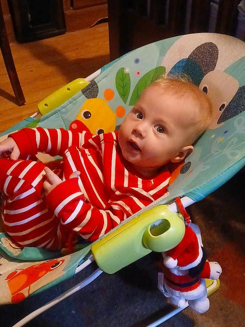 Larry is registered to the contest to win money with this photo: baby, infant, child, striped_pajamas, bouncer, baby_seat, surprised_expression, wide_eyes, indoor, wooden_floor, toy, stuffed_santa, red_white, portrait, head, face, hands, cozy, home, smile